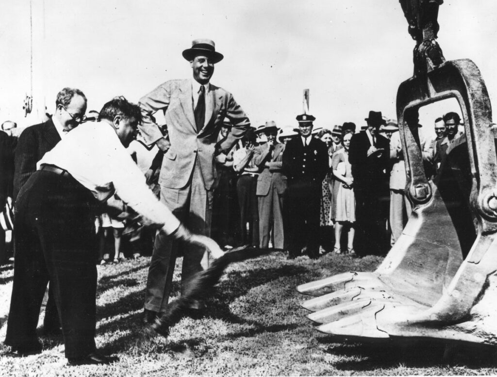 Mayor La Guardia, in white  shirt, lifting the first spade of earth to start construction of New York Municipal Airport. Spectators are in the background. 9-9-1937. (Cradle of Aviation Museum)